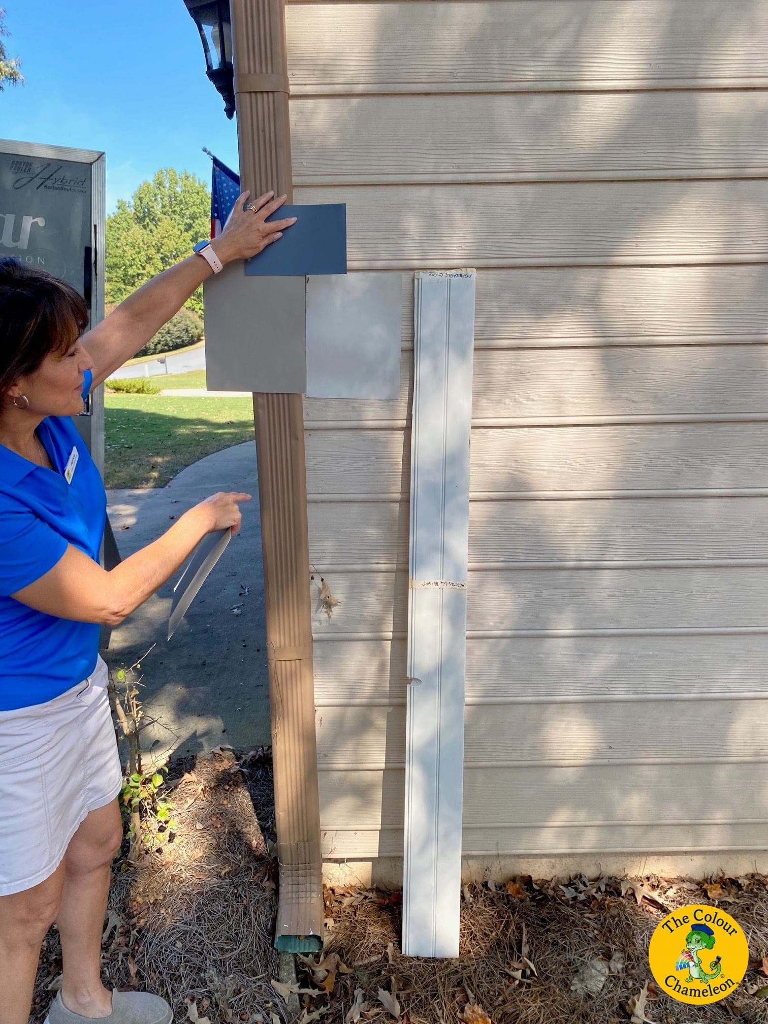 Patricia holding a large blue-grey swatch board against a home's siding beside the downspout, with grey boards and a length of trim propped below during an exterior color consultation