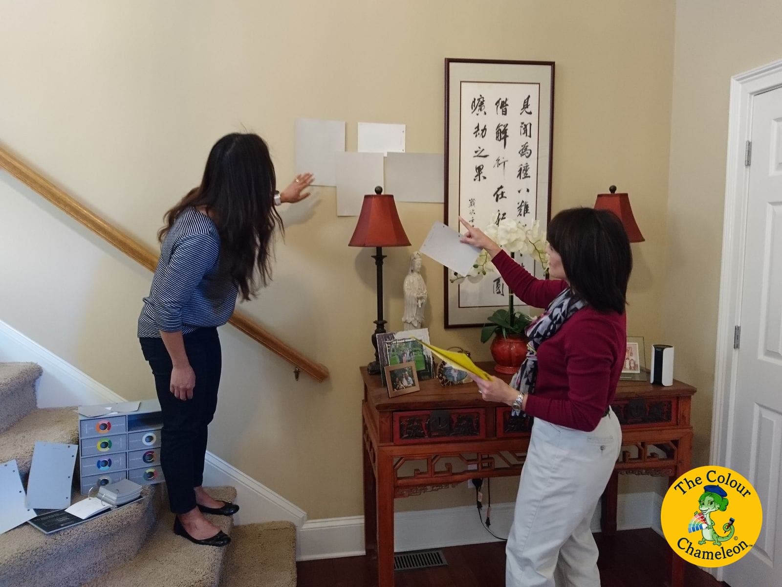 Patricia pointing at large white and grey swatch boards taped to a stairwell wall while a client studies them, with boxes of color decks on the stairs